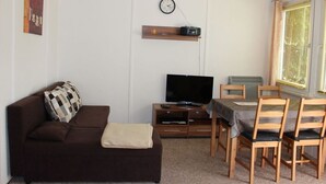 Ein kompakter Wohnraum mit einem braunen Sofa, einem Fernseher auf einem Holztisch, einem Esstisch mit Stühlen und einem Fenster mit Jalousien.