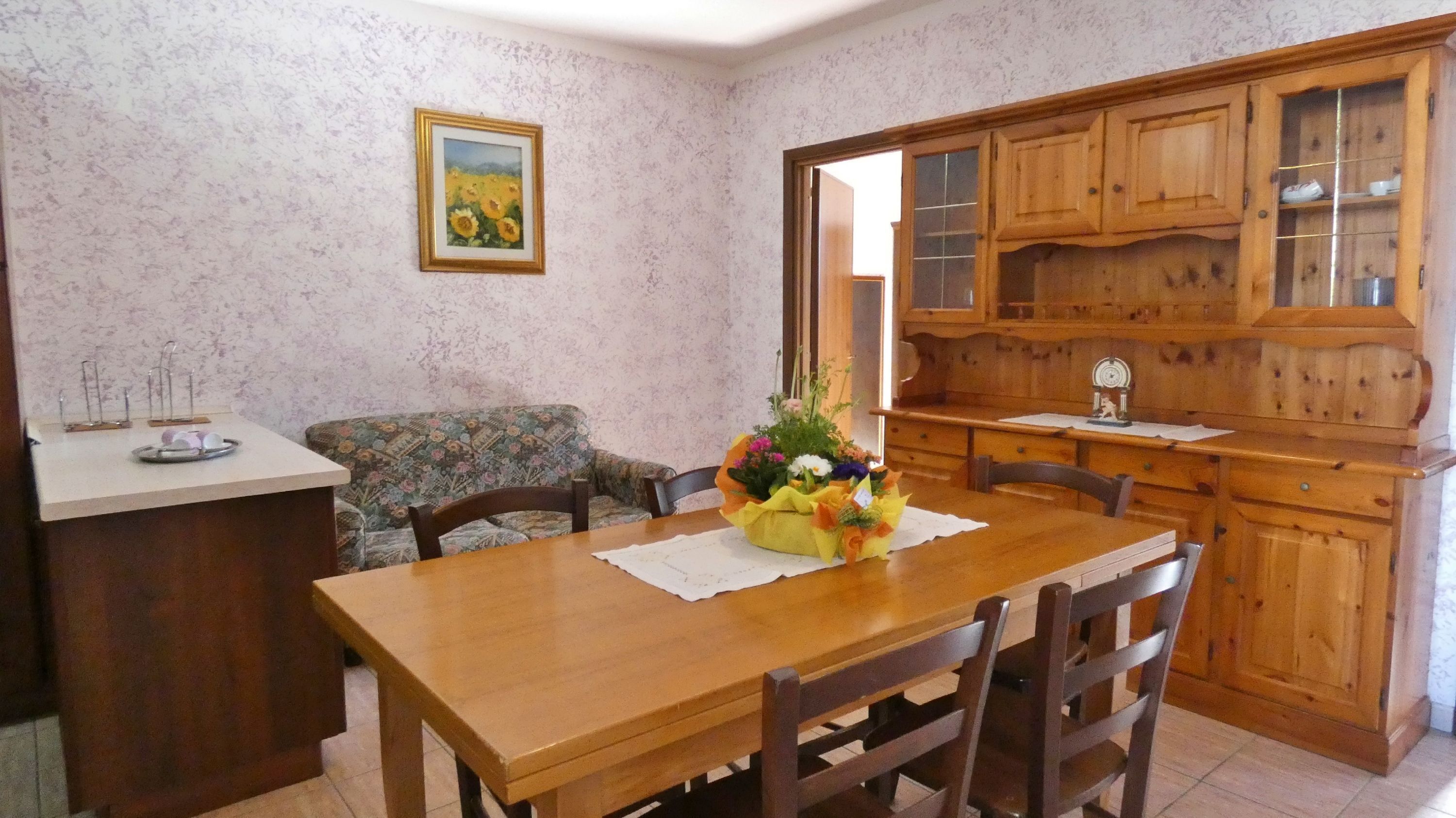 A dining area with a wooden table, chairs, a floral centerpiece, a sofa, and wooden cabinets.