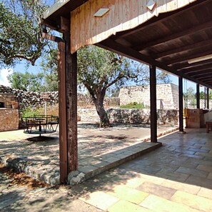 Um pátio externo com piso de pedra, pérgola de madeira e um muro de pedra ao fundo.