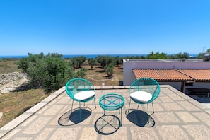 Terraço com duas cadeiras turquesa e uma mesa pequena, com vista para uma paisagem de oliveiras e do mar.