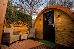 Una cabaña de madera con techo curvo, una puerta negra y mobiliario de mimbre en una terraza de madera.