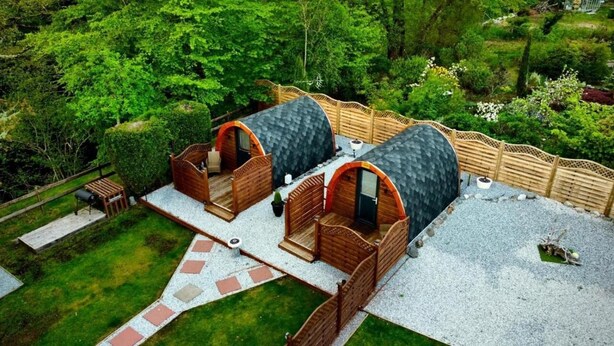 Dos cabañas con techo de paja y interiores de madera, rodeadas por una valla de madera y abundante vegetación.