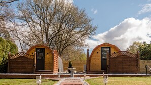 Dos cabañas de madera con un camino de grava que las conecta, rodeadas de árboles y un cielo despejado.