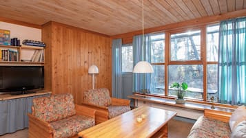 A cozy living room with wooden walls and ceiling, a flat-screen TV, a wooden coffee table, two patterned sofas, a bookshelf, and a large window with curtains.