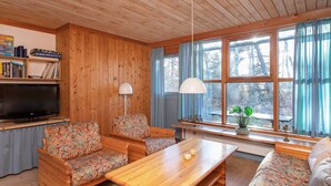 Un salon chaleureux avec des murs et un plafond en bois, un téléviseur à écran plat, une table basse en bois, deux canapés à motifs, une bibliothèque et une grande fenêtre avec des rideaux.