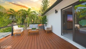 Une terrasse en bois, meublée de meubles en osier, avec une table basse et une vue sur le ciel et la végétation environnante.