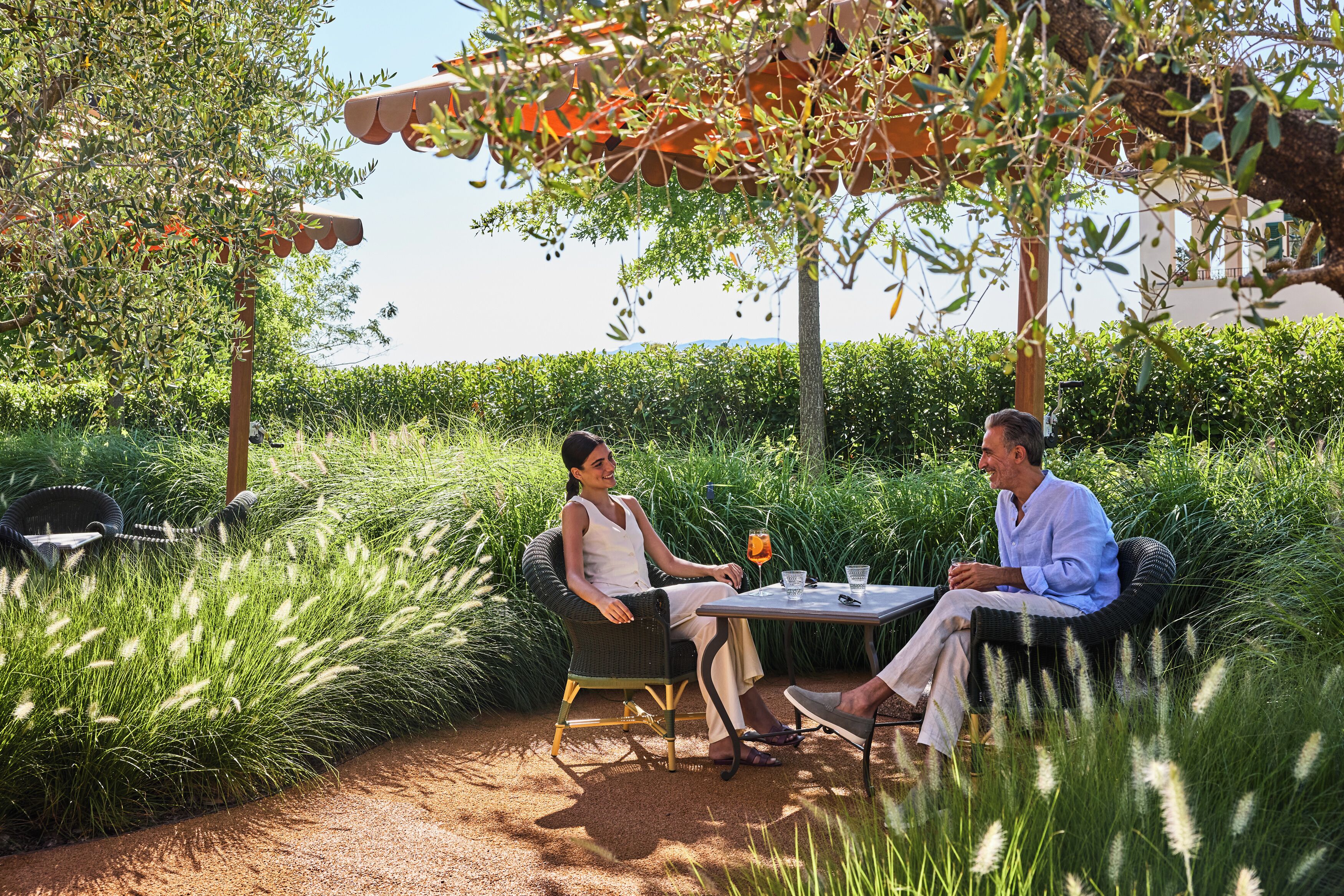 Two people seated at a table outdoors, surrounded by lush greenery and a pergola.