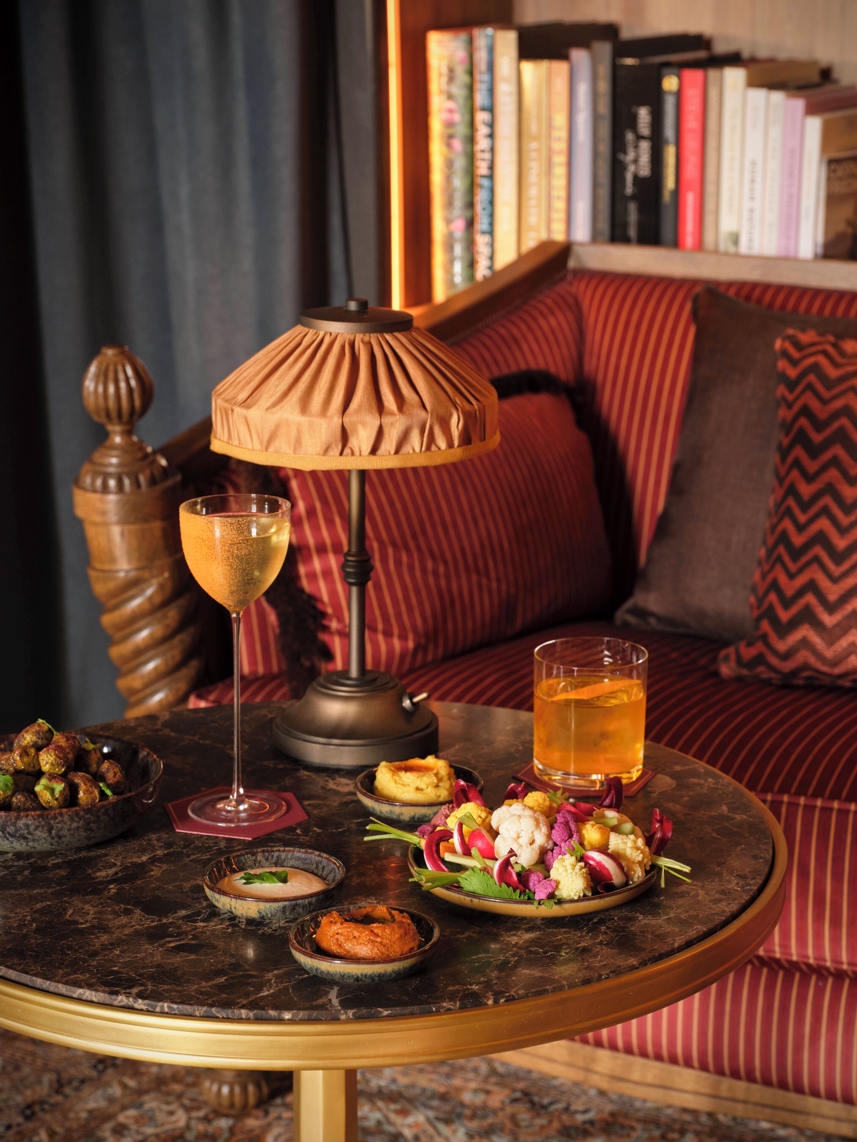 Ein gemütlicher Sitzbereich mit rundem Tisch, einer Lampe, einem Glas bernsteinfarbener Flüssigkeit, einer Schüssel Essen und einem Teller Gemüse.