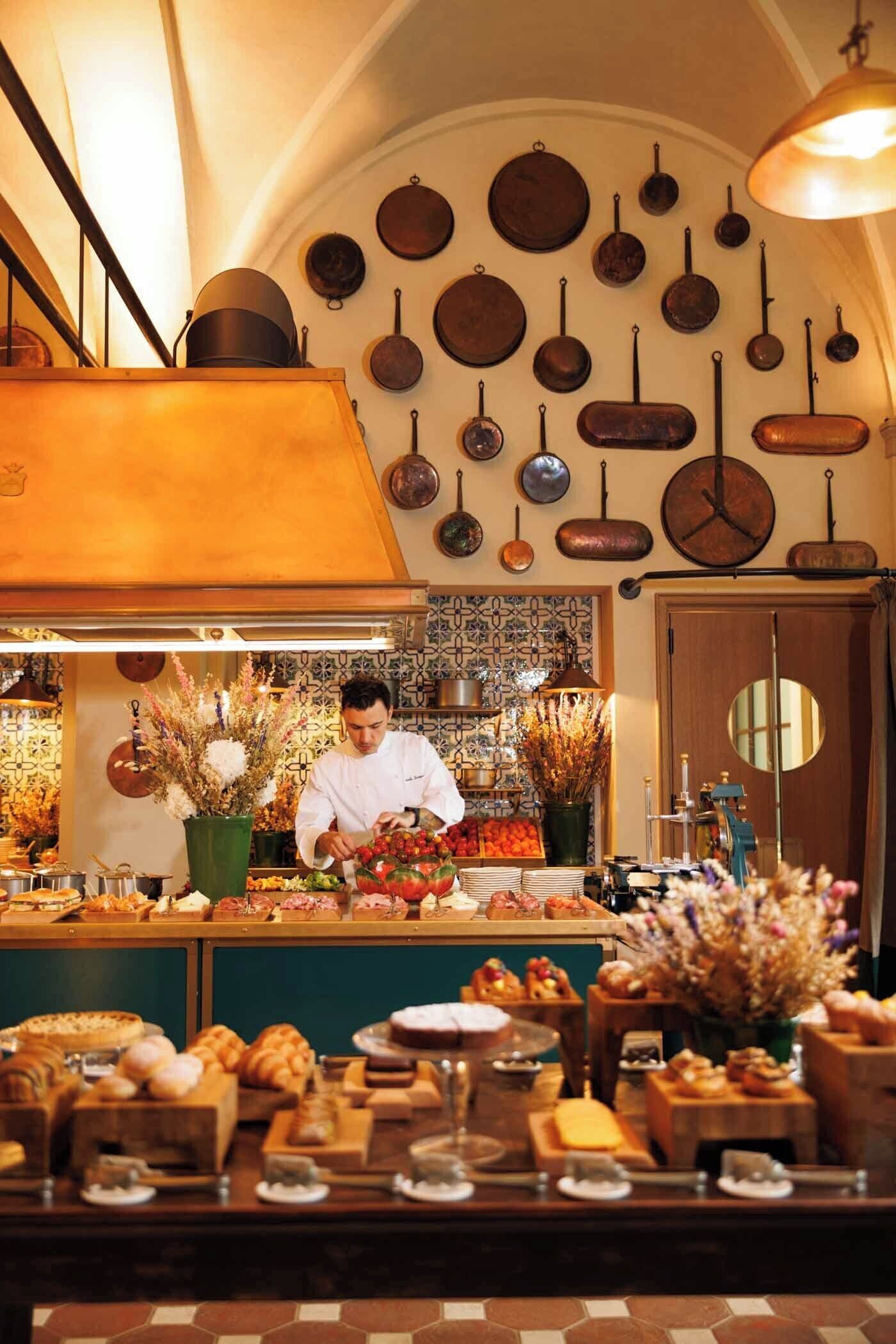 A chef preparing food in a kitchen with hanging pots and a variety of baked goods displayed.