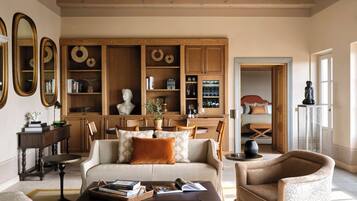 A spacious living room with a sofa, armchairs, and a coffee table. There is a dining area with a table and chairs, and a kitchen area with wooden cabinets and shelves.