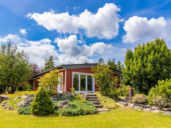 Ein rotes Haus mit Satellitenschüssel, umgeben von üppigem Grün und wolkenlosem, blauem Himmel.