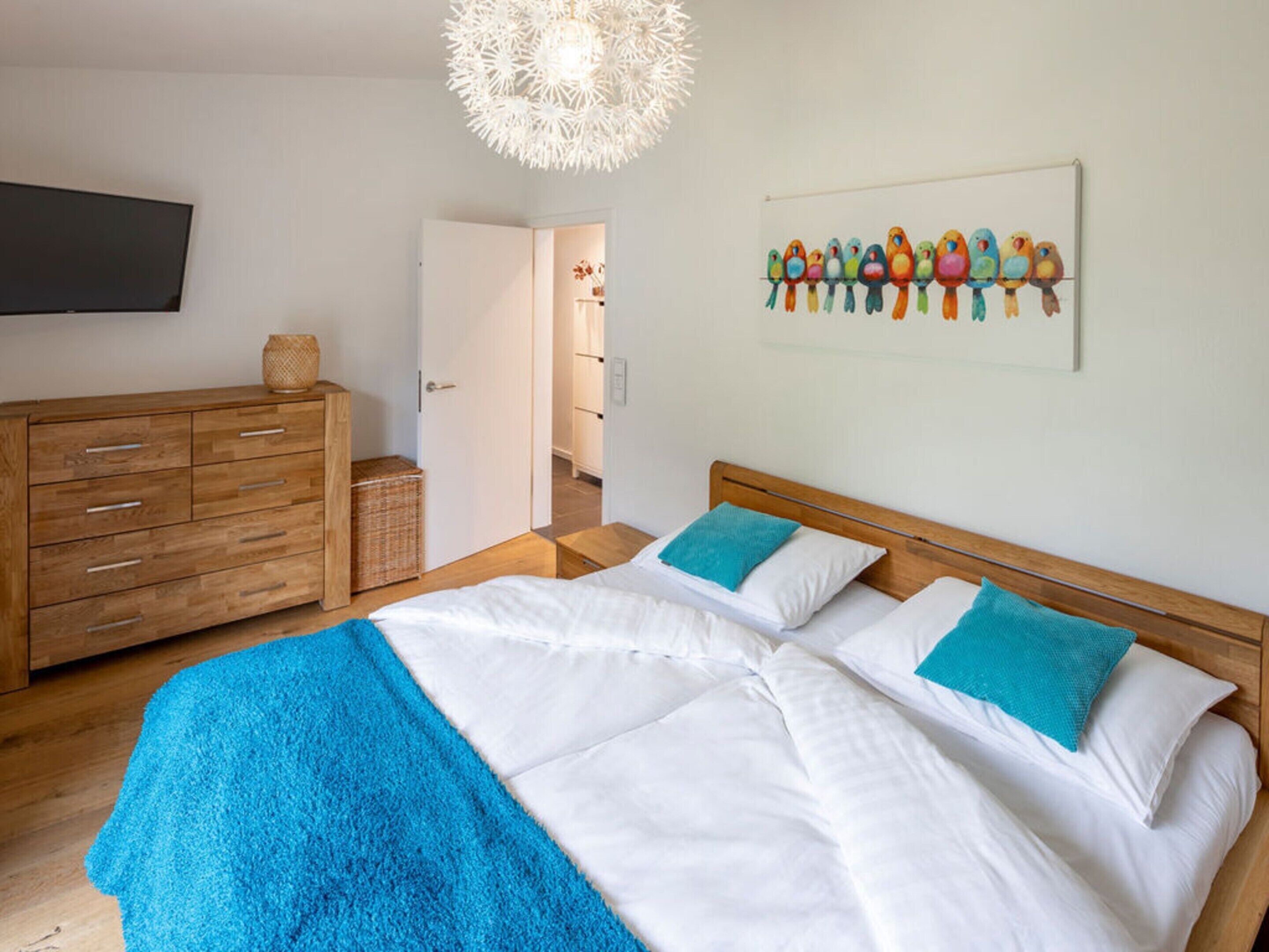 Ein Schlafzimmer mit einem hölzernen Bett, weißer Bettwäsche, einer blauen Tagesdecke, einer hölzernen Kommode, einem Fernseher und einem Wandbild mit Vögeln.