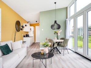 A modern living room with a sofa, coffee table, and dining area with chairs. There is a large window providing a view of the outdoors.