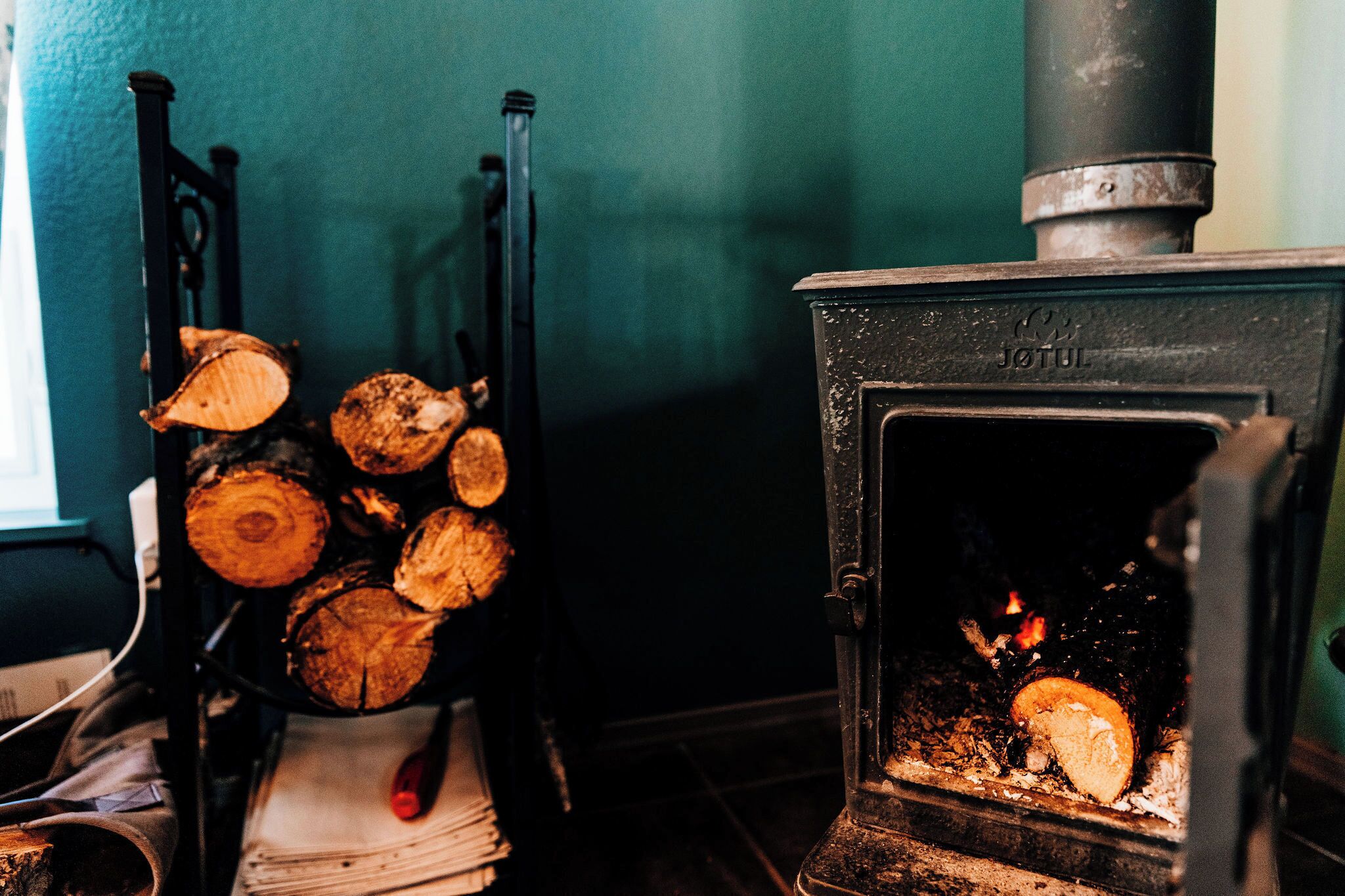 A wood-burning stove with logs and a fire burning inside, a chair with a red cushion, and a window with a view of the outside.