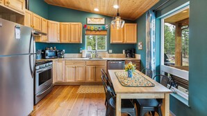 A kitchen with wooden cabinets, a stainless steel refrigerator, a wooden dining table with chairs, and a window with a view of trees. - Sunflower Cabin- Mountain Views- Hot tub (Florissant)