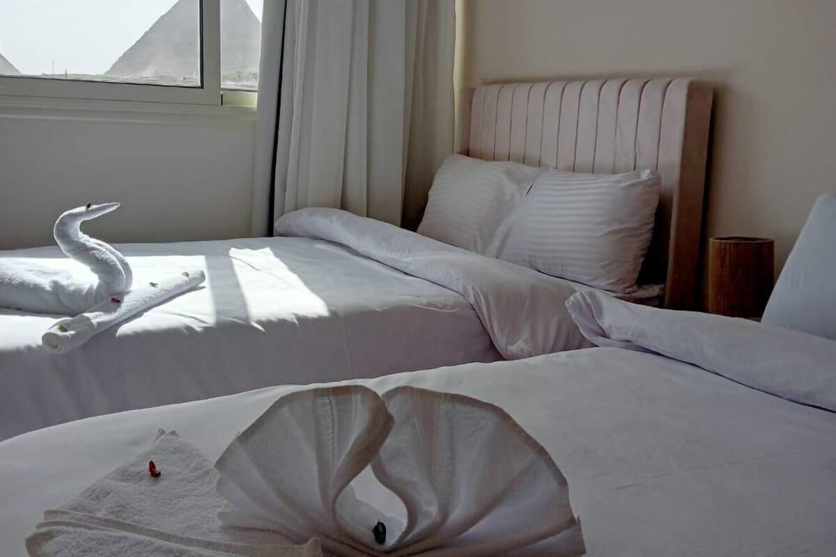 Hotellihuone, jossa on kaksi sänkyä, ikkuna verhoilla, ilmastointilaite ja näkymä pyramideille.