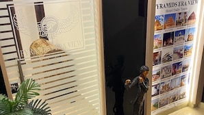 A hotel entrance with a glass door featuring a decorative emblem, a poster with images of tourist attractions, and a statue of a person. - Pyramids Era View (Giza)