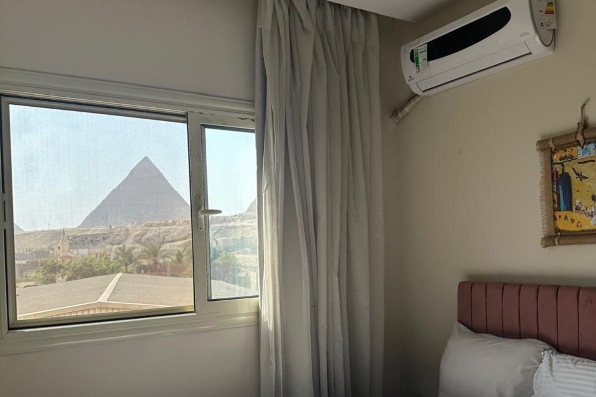 Une chambre d’hôtel avec un lit, une fenêtre donnant sur une pyramide, un appareil de climatisation et un tableau au mur.