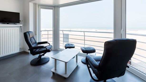 A modern room with a large window overlooking a beach, a flat-screen TV mounted on the wall, and two black leather chairs with a small table in the center. - Apartment on Sea Wall With Stunning Views (Koksijde)