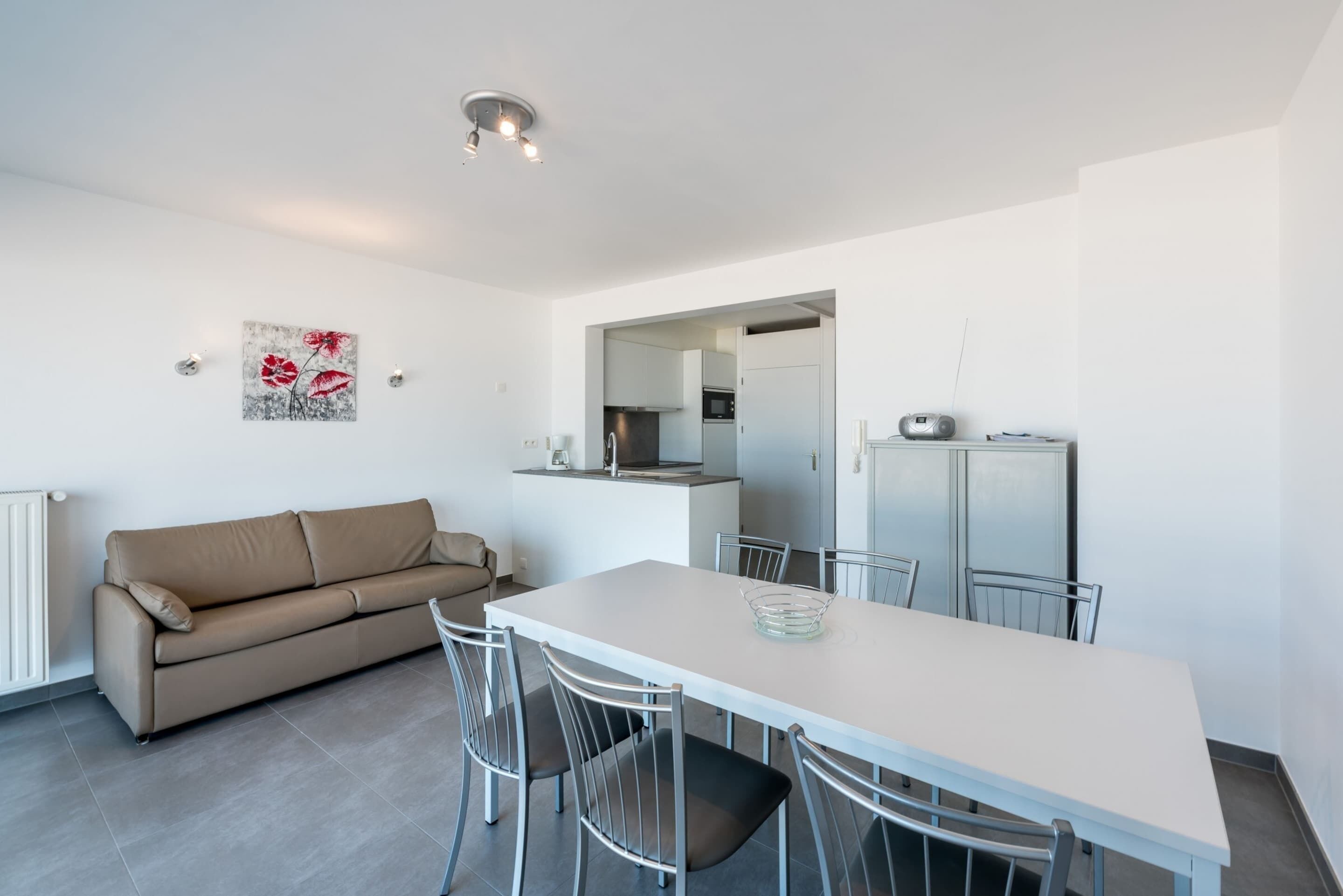 A modern, minimalist living space with a beige sofa, a white dining table with metal chairs, and a kitchen area visible through an open doorway.