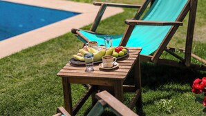 Ein Holztisch mit einer Banane, Äpfeln, einem Glas Wasser und einer Kaffeetasse auf einer Rasenfläche in der Nähe eines Pools.