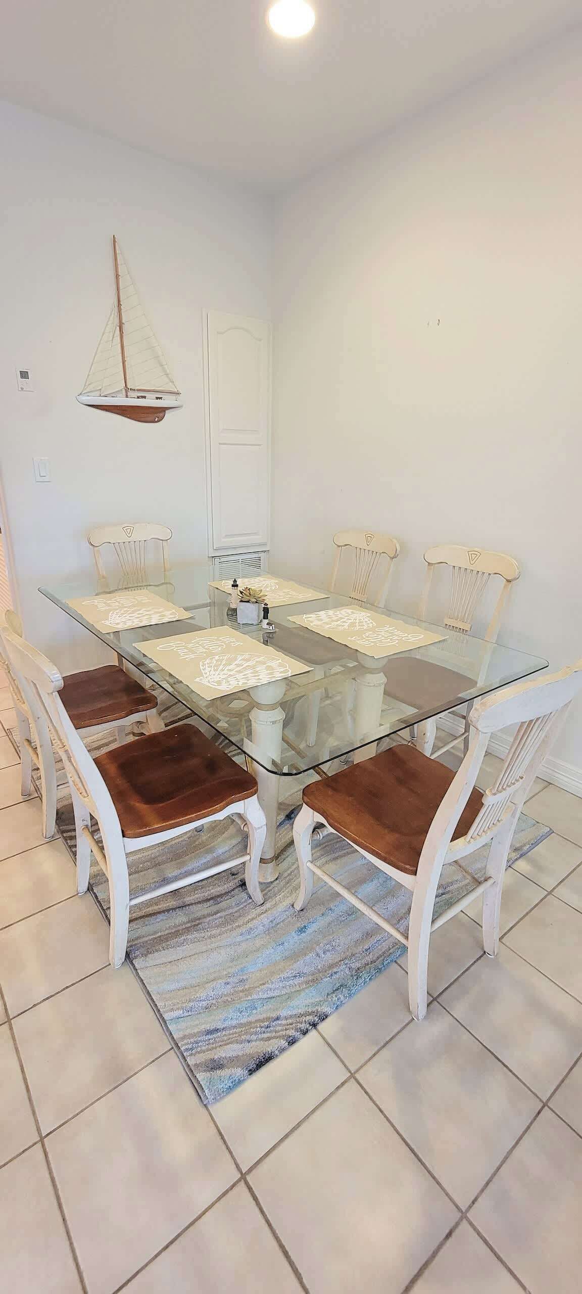 Un coin repas avec une table à plateau de verre, des chaises blanches avec des coussins marron, un tapis à rayures et une décoration représentant un voilier sur le mur.