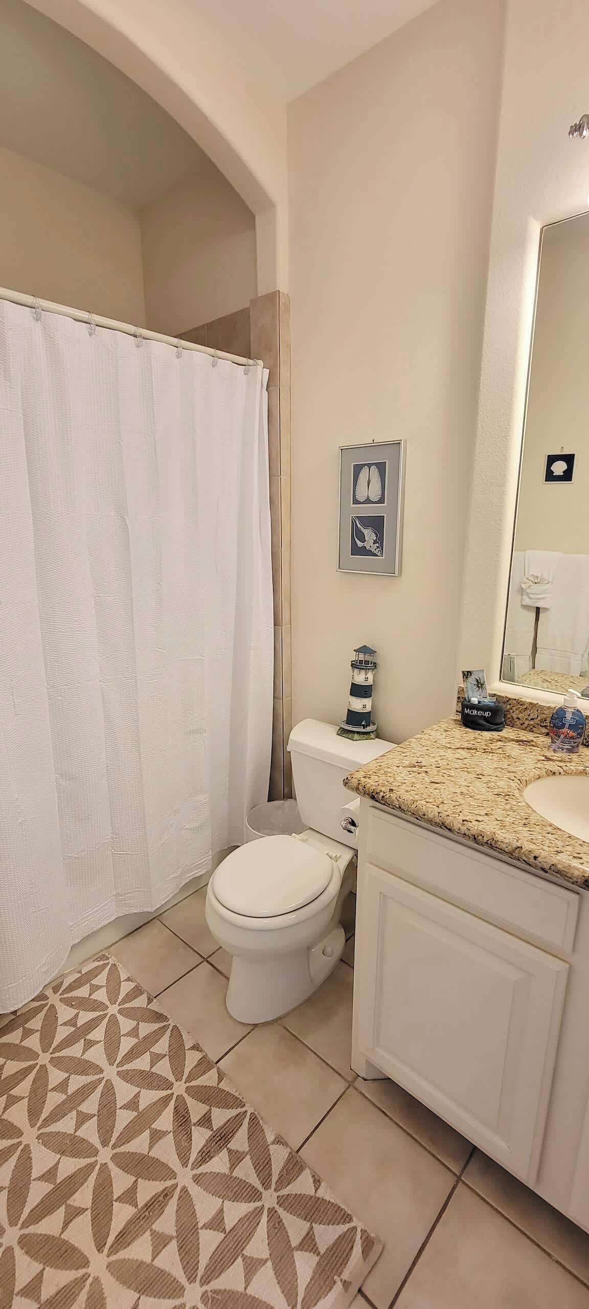 A bathroom with a toilet, sink, and shower curtain. There is a rug on the floor and a small lighthouse decoration on the wall.