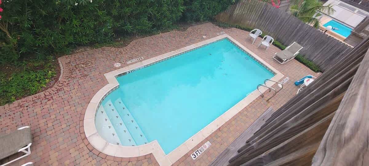 Una piscina rettangolare con bordo in mattoni, circondata da una recinzione in legno e una piccola zona con sedie.