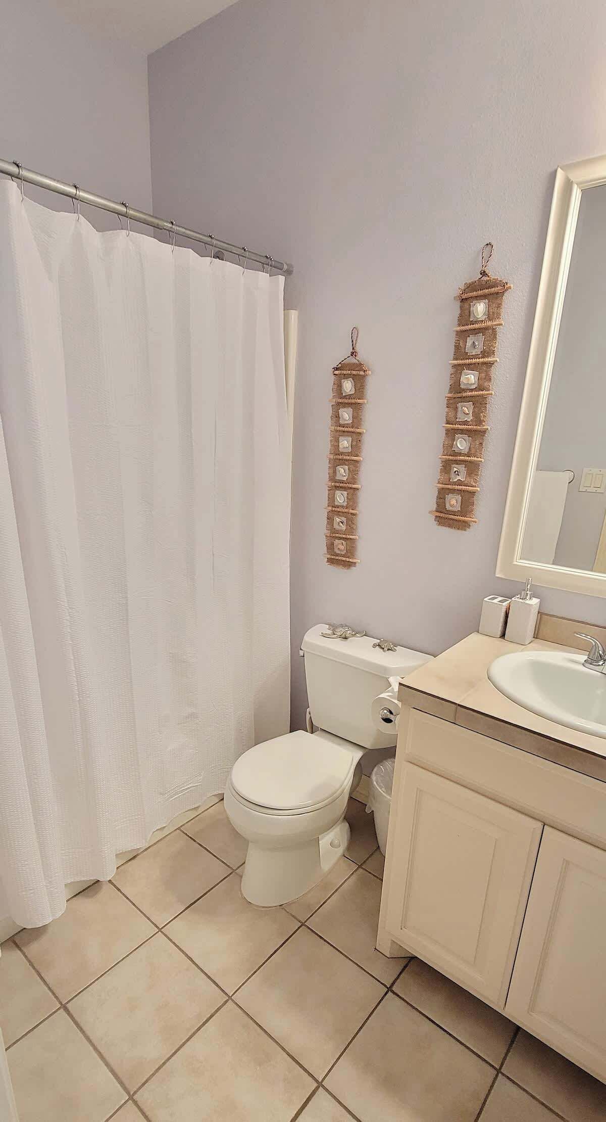 A bathroom with a toilet, sink, and mirror. There is a shower curtain and a wall-mounted shelf with decorative items.