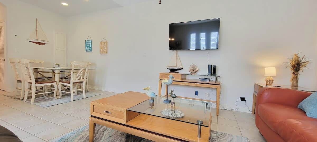 A living room with a wooden coffee table, a glass-top side table, a red sofa, a TV mounted on the wall, and a dining area with a white table and chairs.