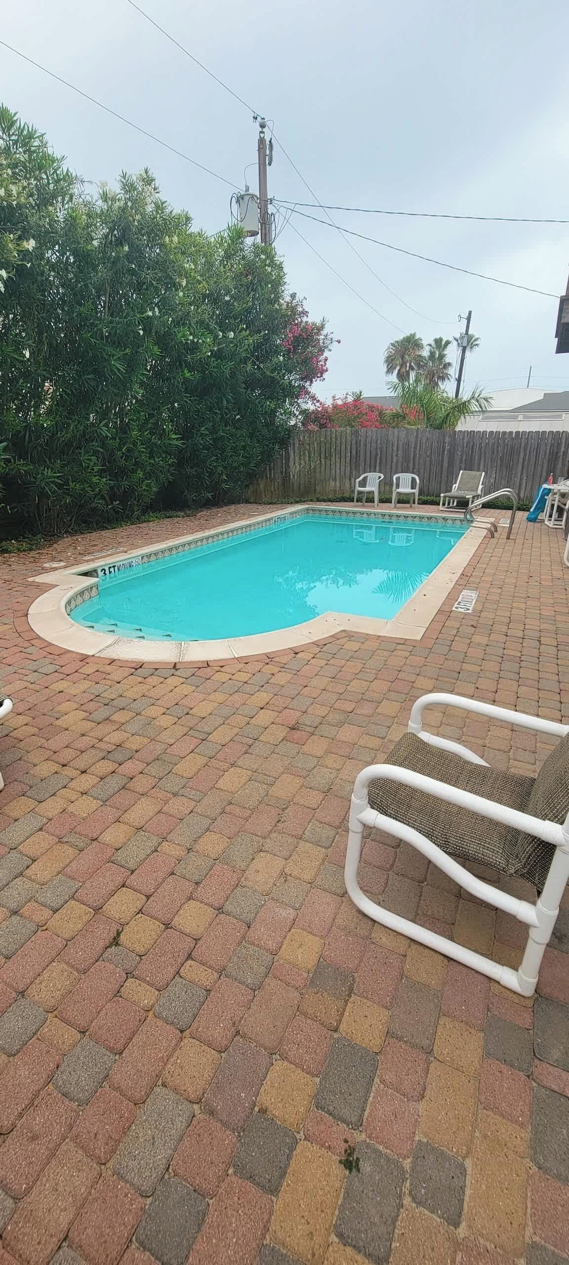 A swimming pool with a tiled deck, surrounded by a wooden fence and greenery.