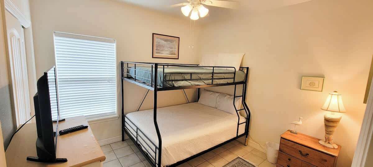 A bunk bed with a desk and a TV, a window with blinds, a ceiling fan, and a lamp on a dresser.