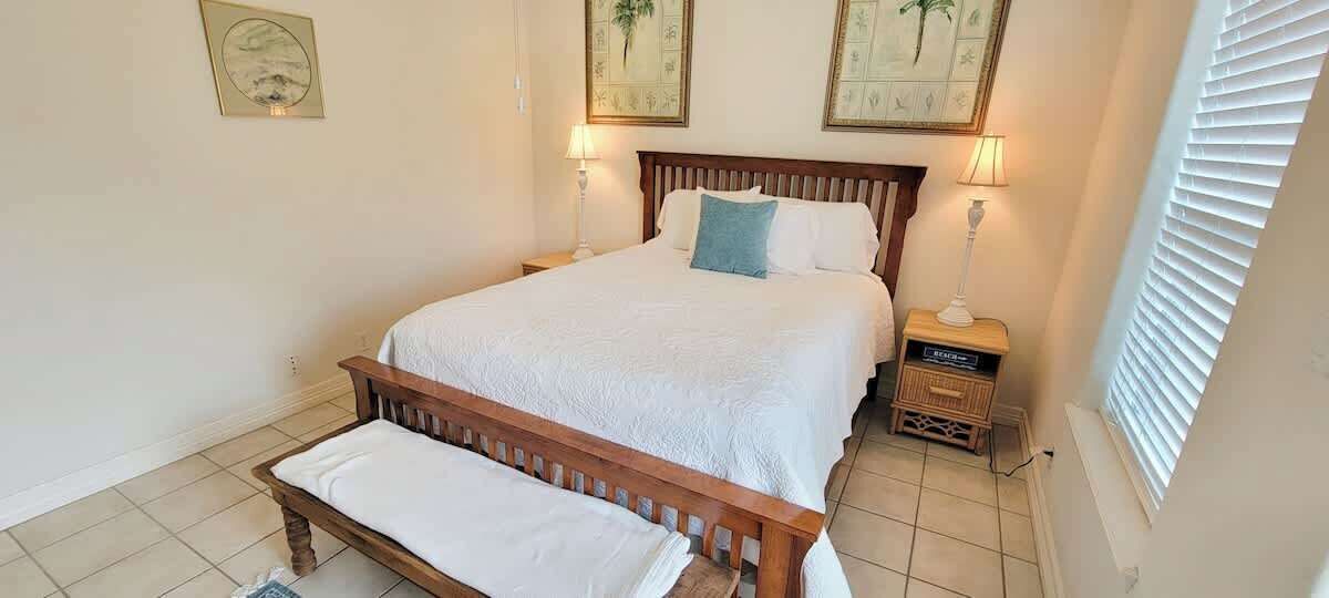 Une chambre à coucher avec un lit en bois, du linge de lit blanc, deux lampes de chevet et une fenêtre avec des stores.