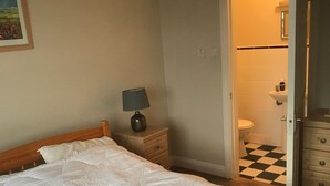 Une chambre avec un lit en bois, une table de chevet avec une lampe, une salle de bain visible à travers une porte ouverte et un sol à carreaux.