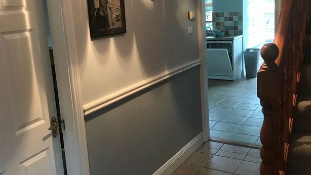 Un couloir avec une porte blanche, un tableau encadré accroché au mur et une cuisine visible à travers une porte ouverte.
