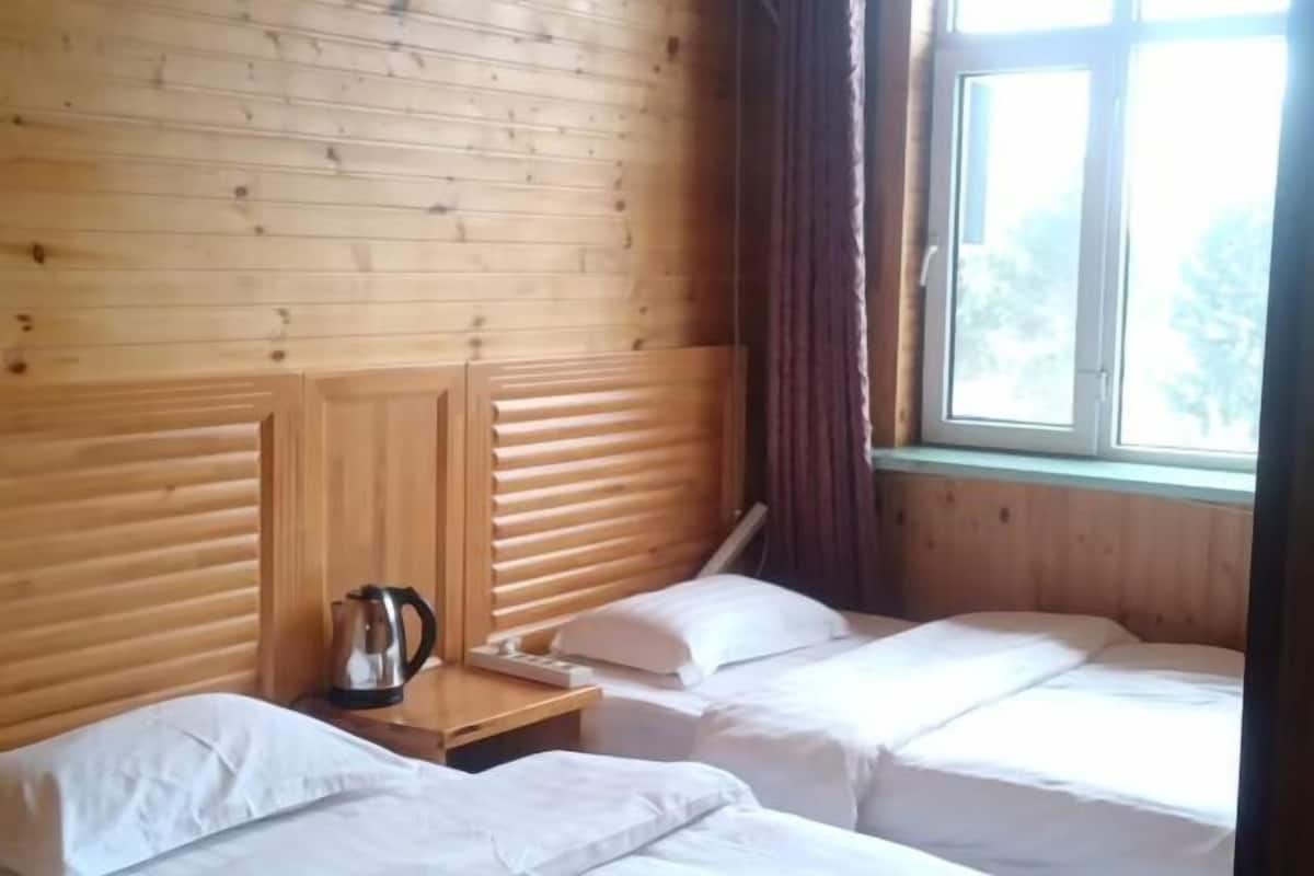 Quarto com painéis de madeira, duas camas com lençóis brancos, uma mesinha com chaleira e uma janela com cortinas.