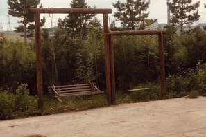 A concrete pathway with a wooden swing set and a bench in a wooded area.
