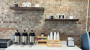 A coffee station with a brick wall background, coffee makers, cups, and a sign that says "Now Serving." - The Hudson Inn (Morris)