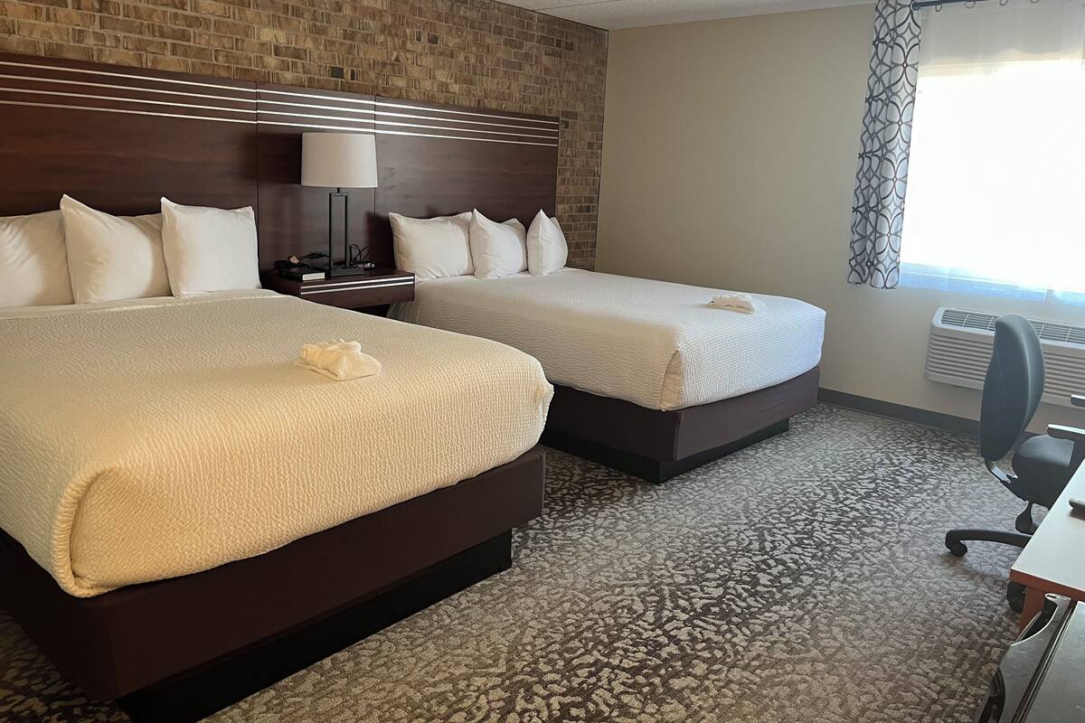 A hotel room with two beds, a brick wall, a patterned carpet, a desk, and a window with a curtain.