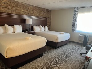 A hotel room with two beds, a brick wall, a patterned carpet, a desk, and a window with a curtain.