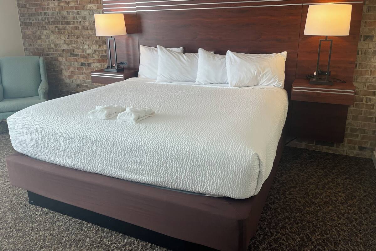A hotel room with a large bed, two bedside lamps, a brick wall, and a blue armchair.
