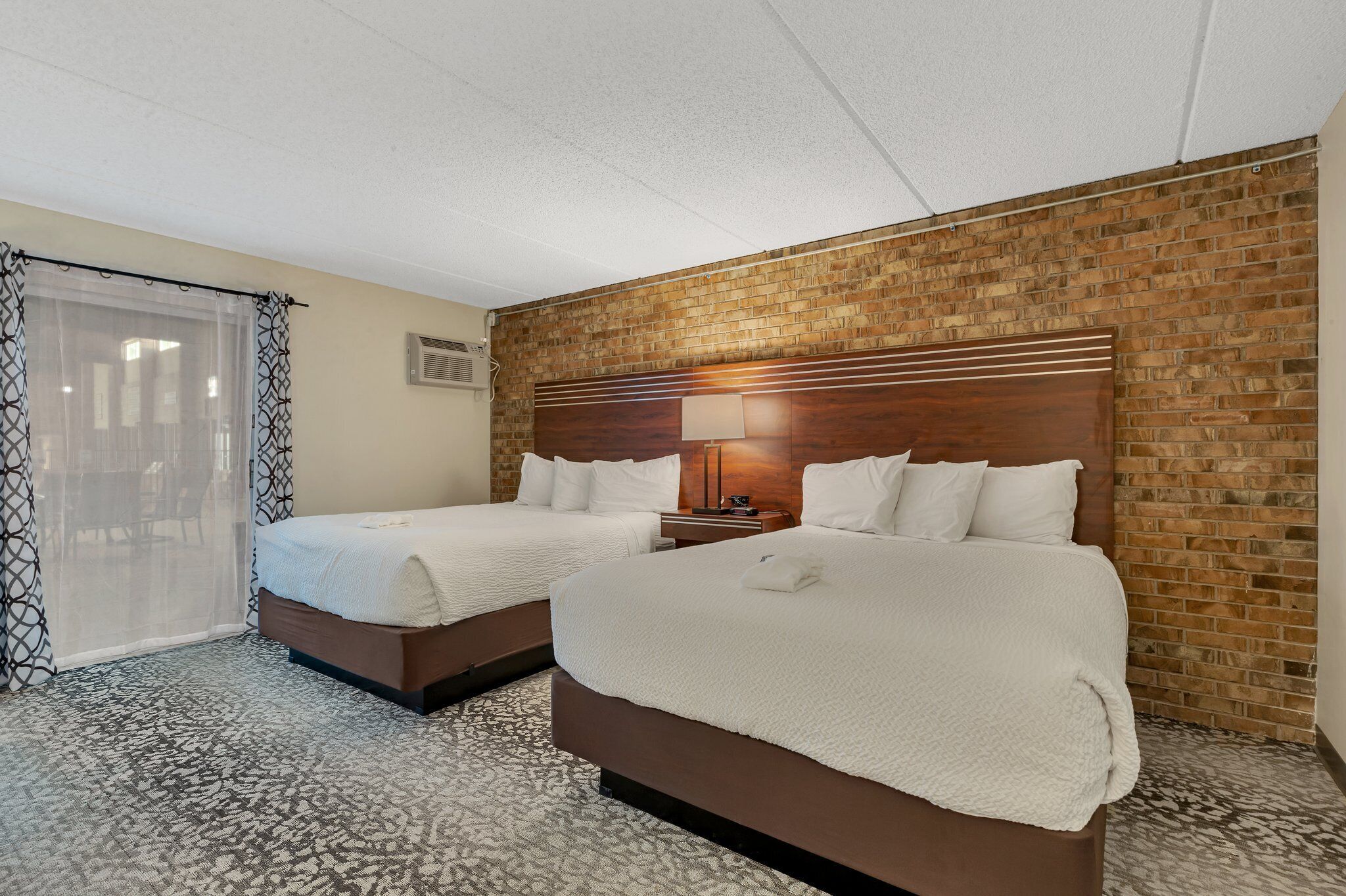 A hotel room with two beds, a brick accent wall, a patterned carpet, and a window with sheer curtains.