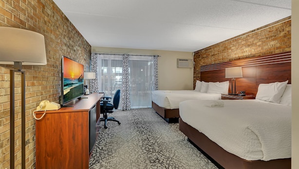 A hotel room with a brick wall, a desk with a telephone, a chair, a bed with white bedding, and a window with sheer curtains. - The Hudson Inn (Morris)