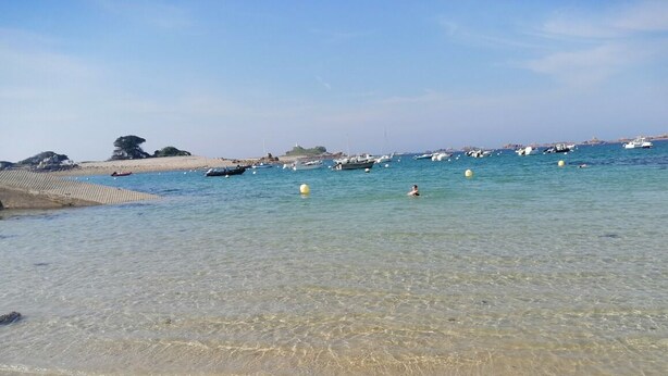 A beach with clear shallow waters, boats in the distance, and a sandy shore.