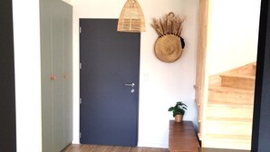 A hallway with a dark door, a woven light fixture, a woven wall hanging, a small wooden bench with a potted plant, and a wooden staircase.