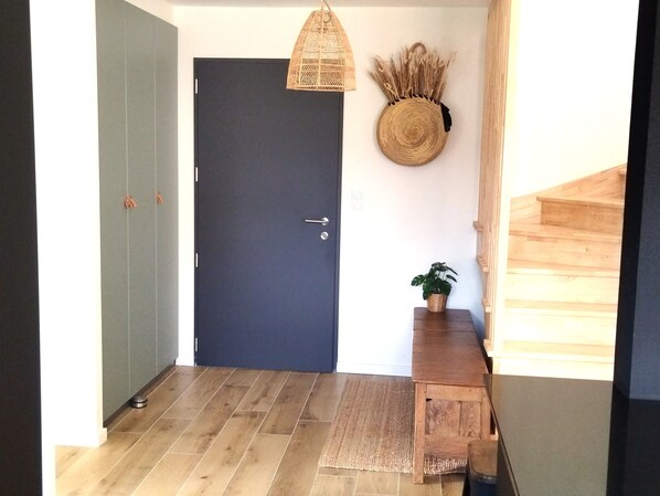 Ein Flur mit einer dunklen Tür, einer gewobenen Leuchte, einem Wandbehang aus Gewebe, einer kleinen Holzbank mit einem Topf und einer Holztreppe.
