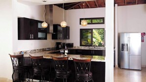 A modern kitchen with dark cabinetry, a marble island with bar stools, pendant lighting, and a stainless steel refrigerator.