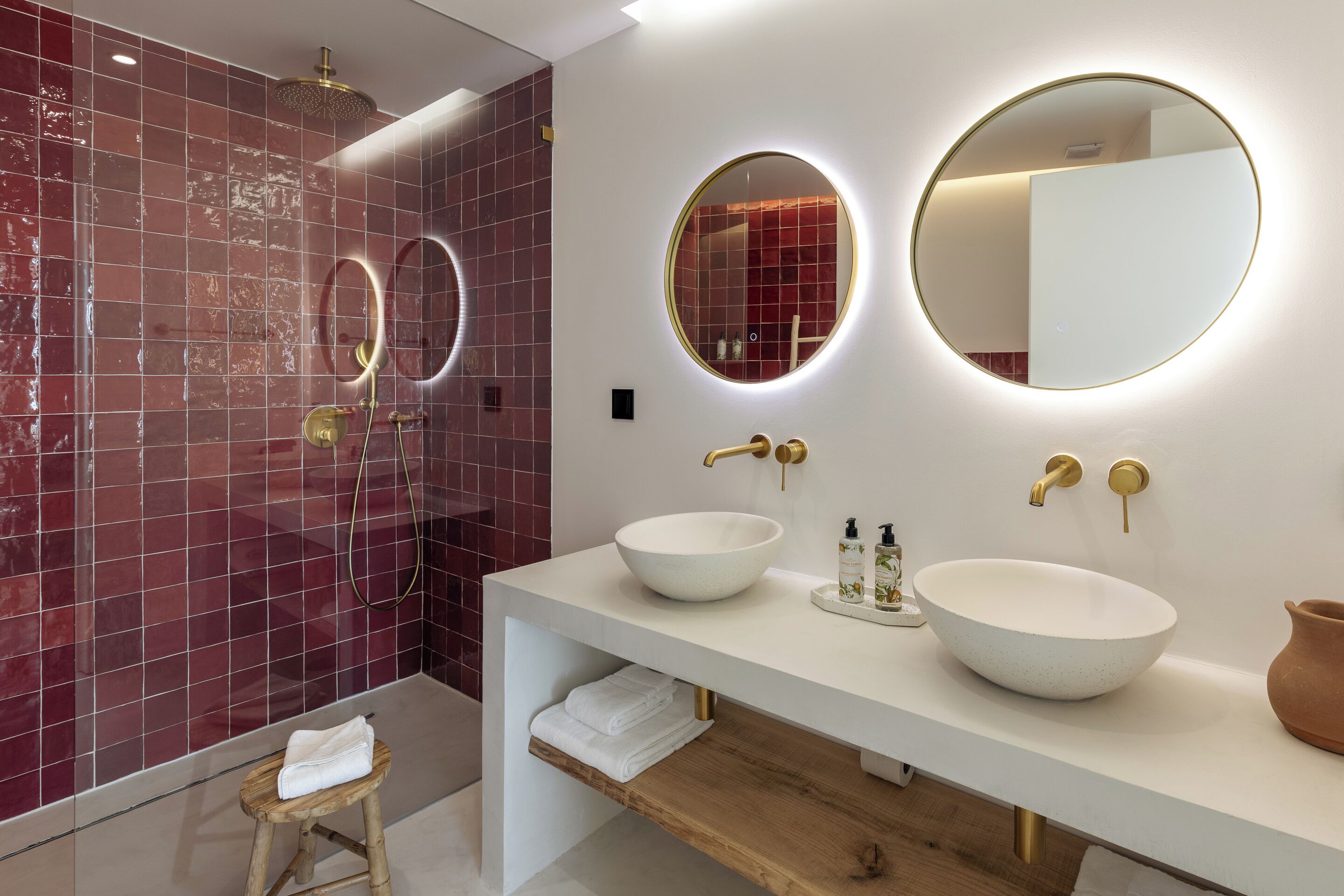 Casa de banho moderna com lavatório branco, duas espelhas redondas, uma grande pia branca e um chuveiro com parede em azulejos vermelhos.