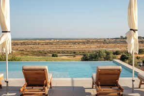 Zona de piscina con dos sillones de madera y sombrillas blancas, con vistas a un paisaje extenso.