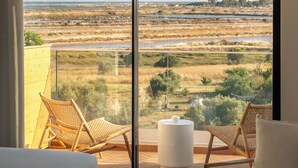 A room with a large sliding glass door leading to a balcony with a view of a coastal landscape. There are two wicker chairs and a white ottoman on the balcony. - Villa Loreto (Olhao)
